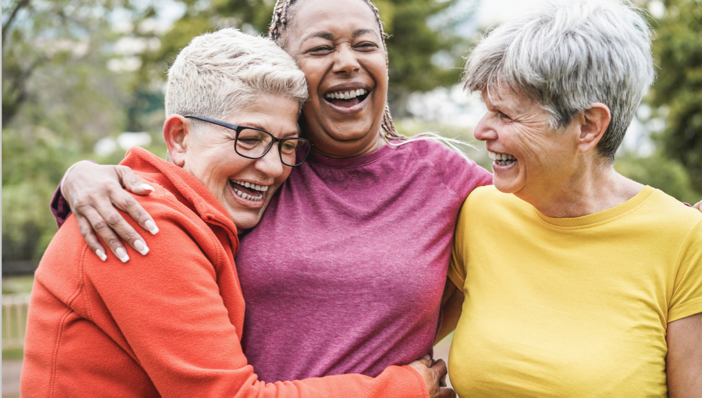 Three older women outside in workout clothes, laughing with their arms around one another