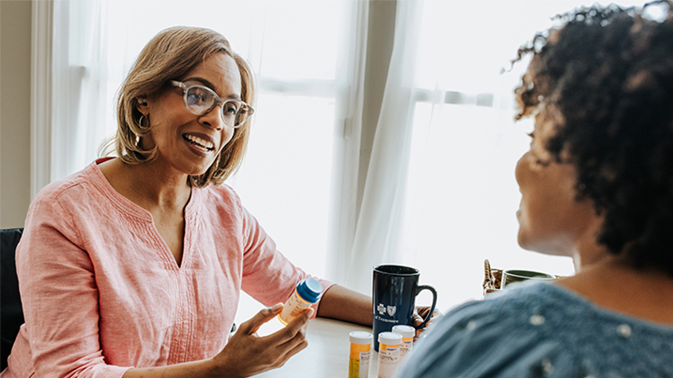 Woman holding a medication bottle and talking to her daughter at the kitchen table.