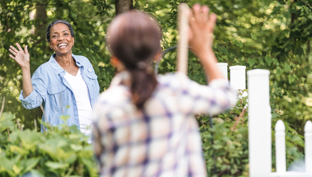 Two women waving at each other across their shared garden fence.
