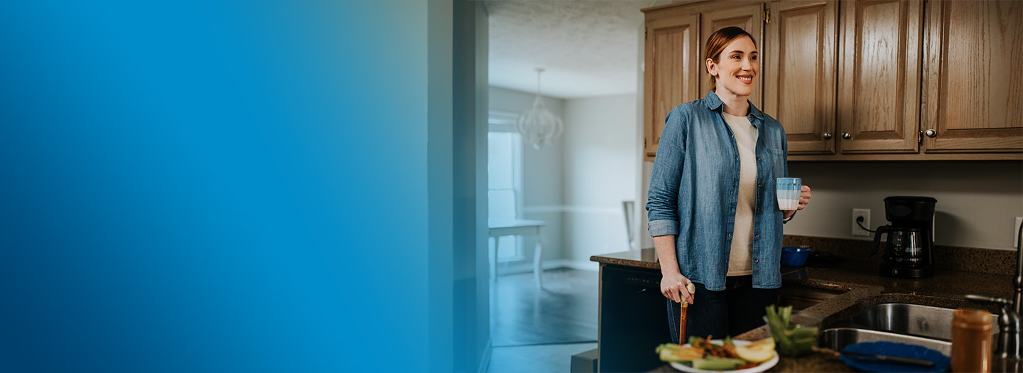 A woman with a cane standing in her kitchen and enjoying coffee.